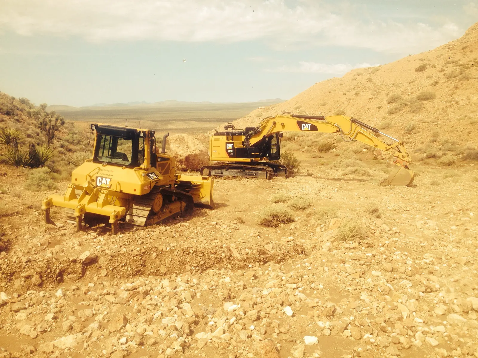 A CAT D6 dozer with a ripper and a CAT 320E excavator on a rocky desert site among Joshua trees