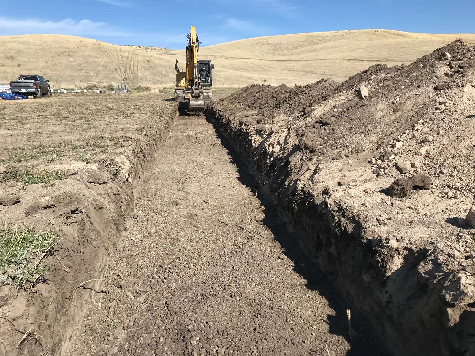 An excavator at the end of a long, wide trench cut through a pasture, with dry hills behind