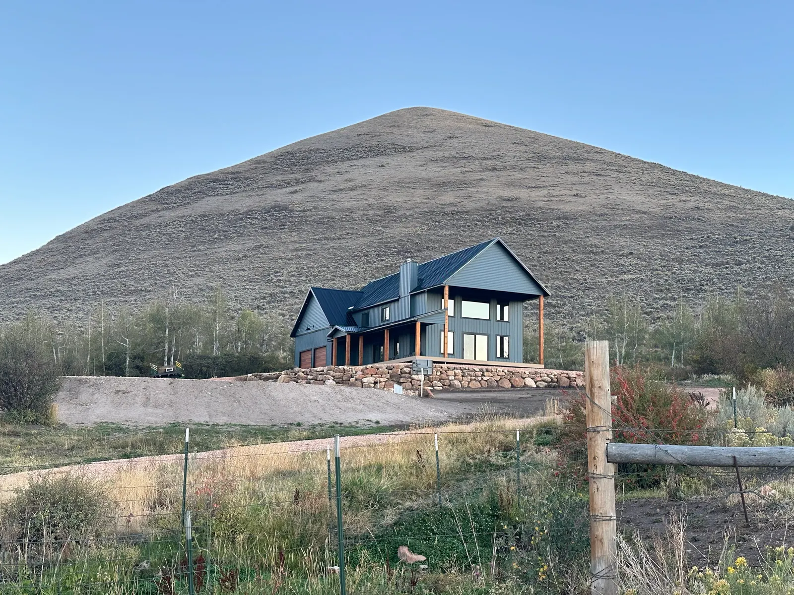 A new two-story home with gray board-and-batten siding, a black metal roof and cedar porch posts, set on a graded lot above a boulder retaining wall, with a sagebrush hill behind it