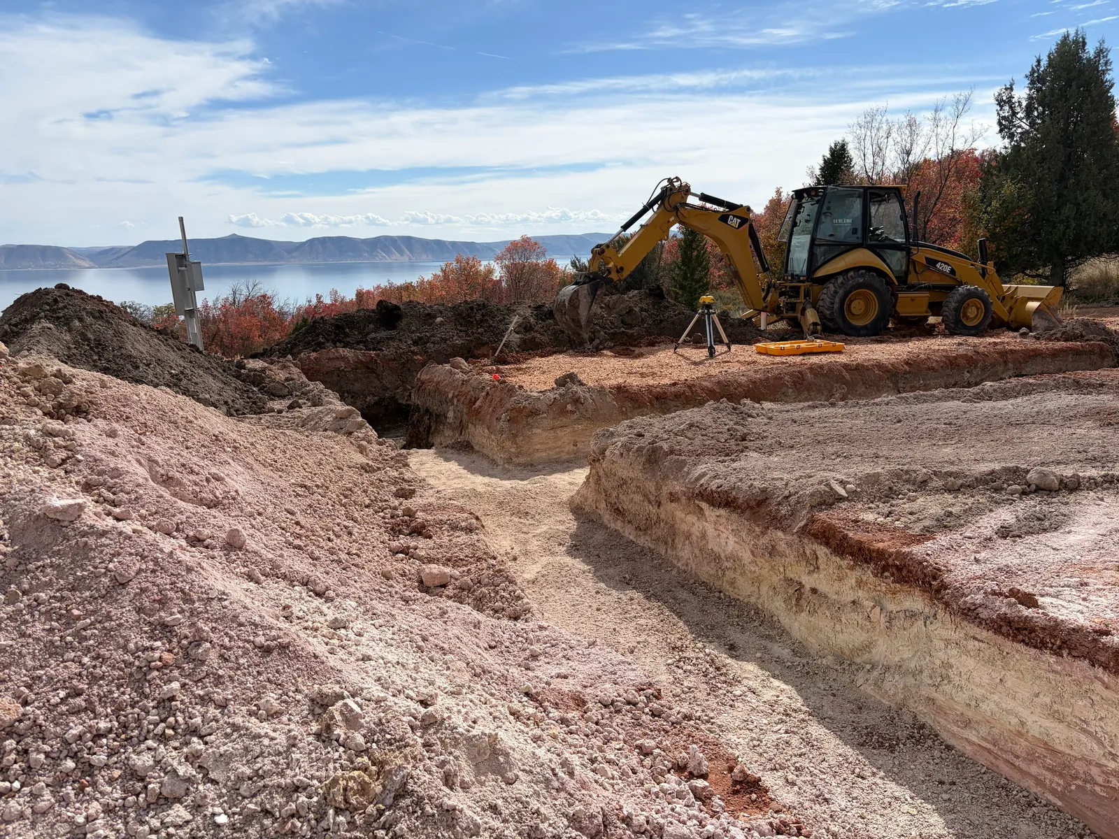 A foundation excavation cut to depth on a hillside above Bear Lake, with a CAT backhoe and a laser level beside it