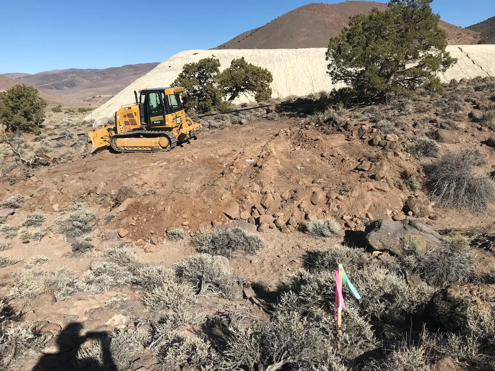 A CAT dozer clearing rocky, sagebrush-covered ground below a hillside, with survey flags in the foreground