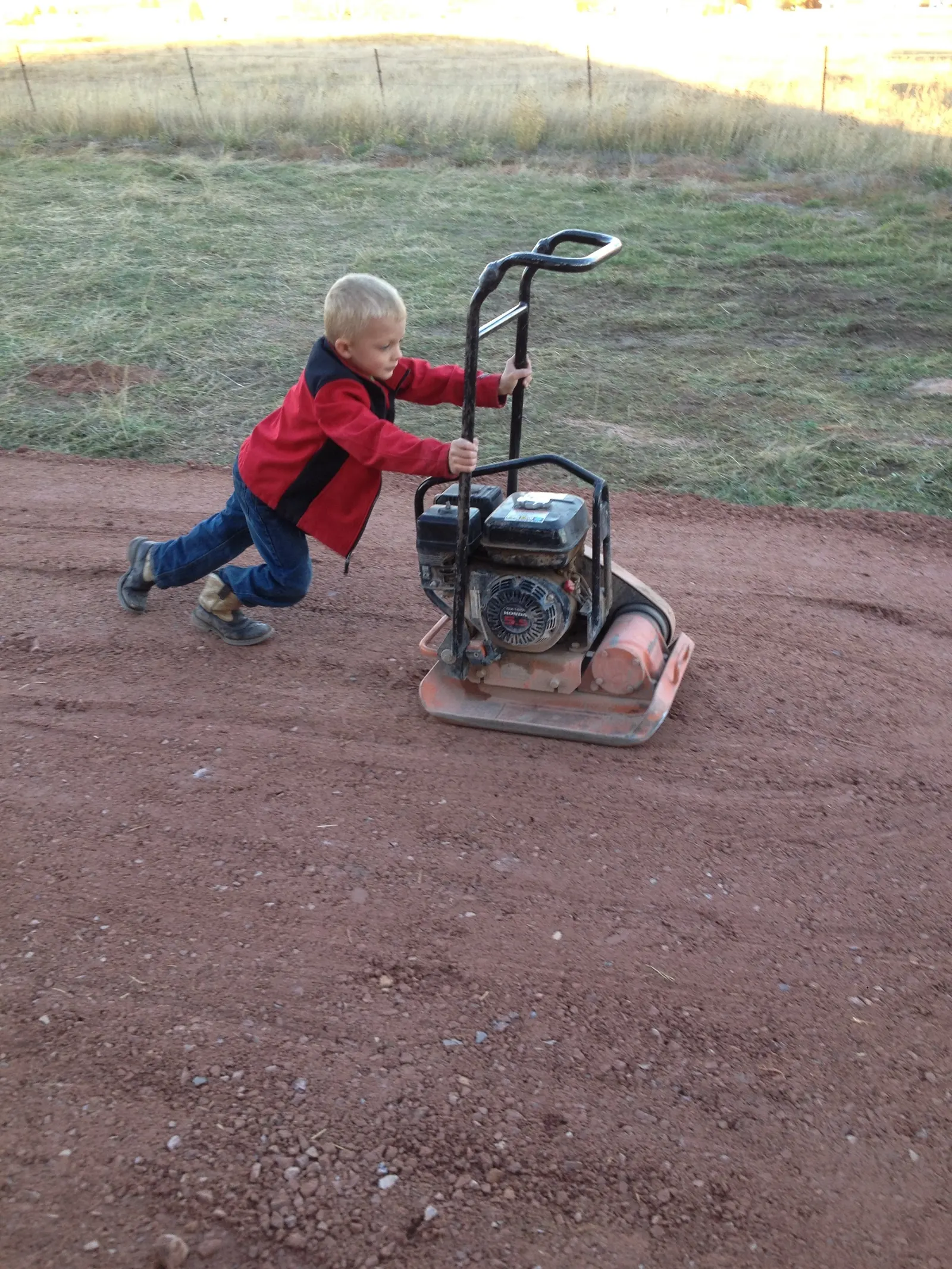 A young boy pushing a plate compactor across a freshly graded red gravel driveway