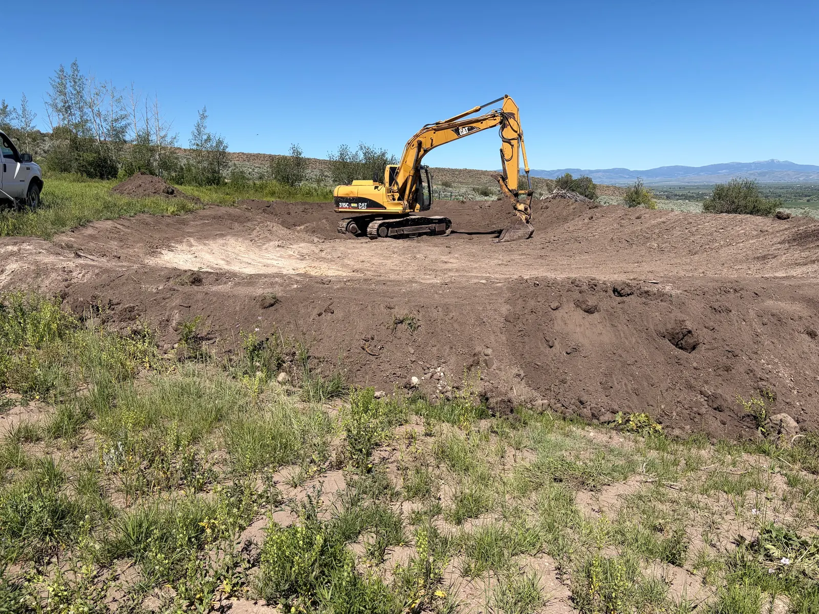 A CAT 315C excavator shaping a large earthen basin on a hillside, with the valley and mountains beyond