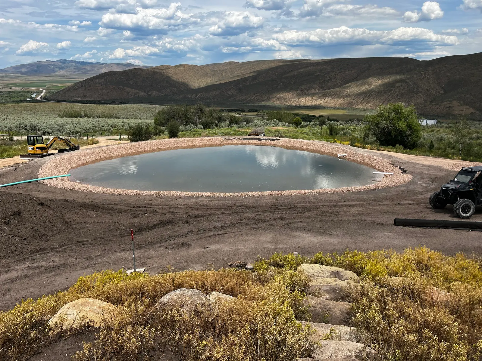 A finished, filled pond ringed with gravel on a hillside, with sagebrush, a valley and mountains beyond under a cloudy sky