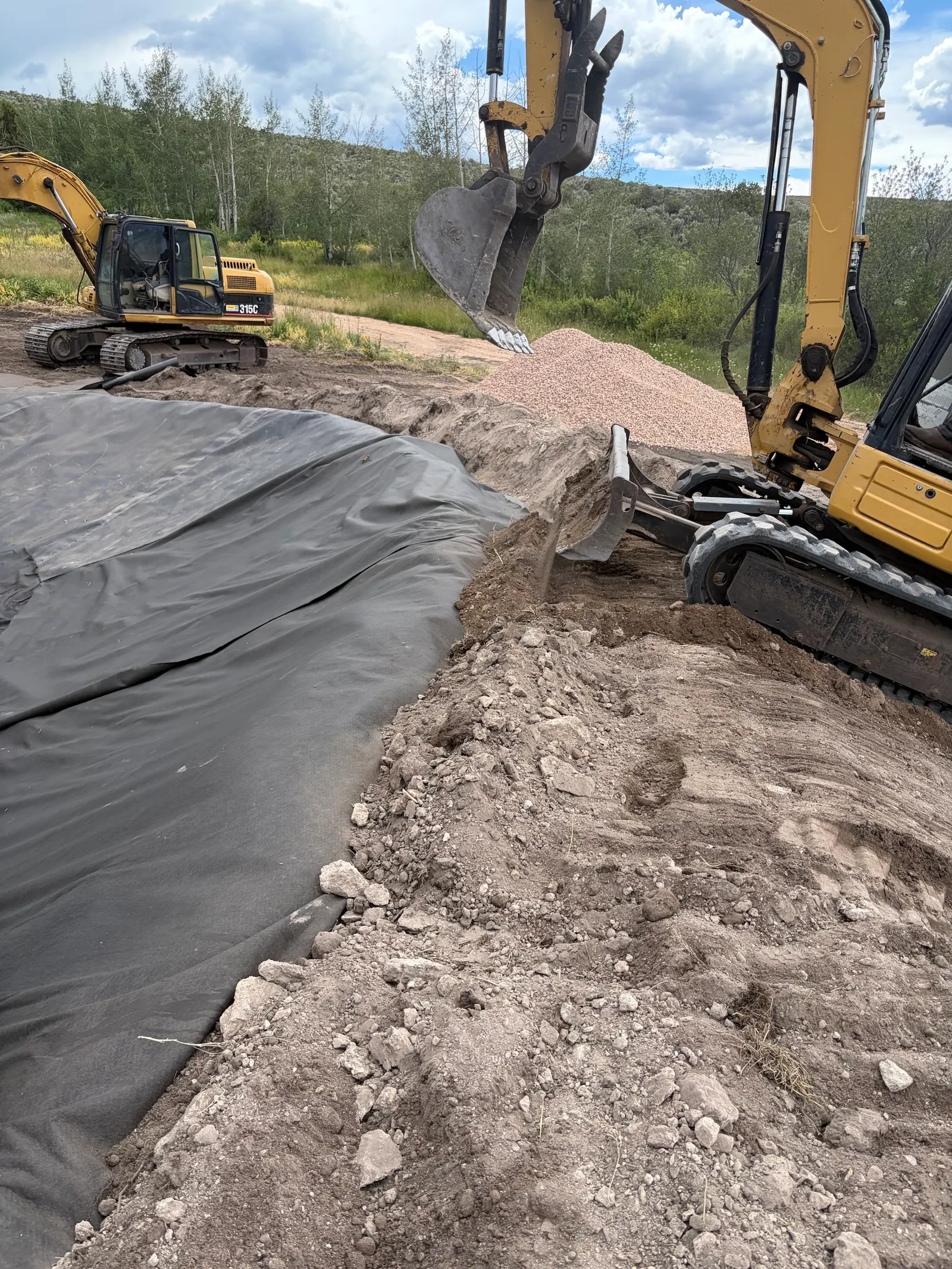 A mini excavator pushing soil over the edge of a black liner, with a larger CAT 315C excavator and a pile of gravel behind