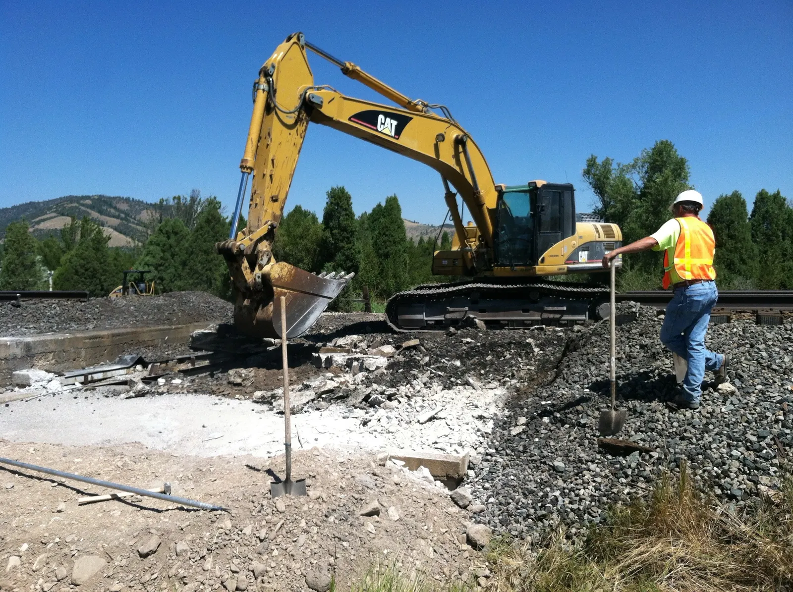 A CAT excavator breaking out old concrete beside a railroad track while a crew member in a hard hat and safety vest stands by
