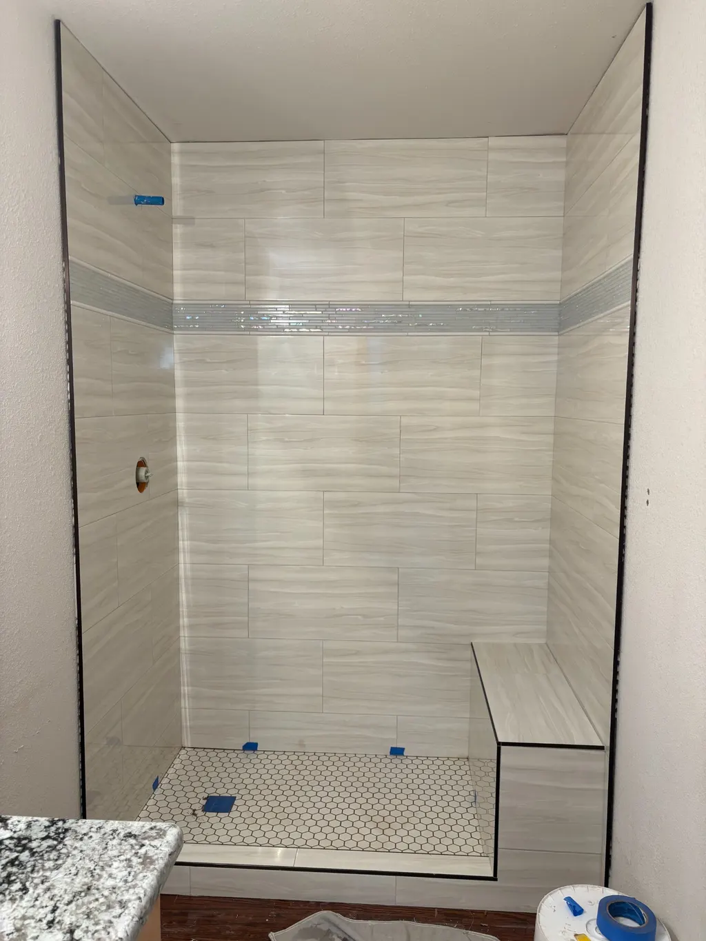 A newly tiled walk-in shower with large-format plank tile, an iridescent glass accent band, a built-in bench and a white hexagon floor
